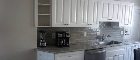 A kitchen with white cabinets, a granite countertop, a coffee maker, a blender, and a dishwasher.
