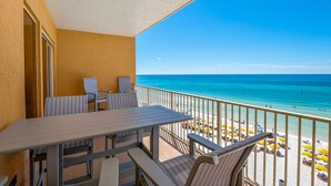 Un balcone con tavolo e sedie, affacciato su una spiaggia con ombrelloni e persone.
