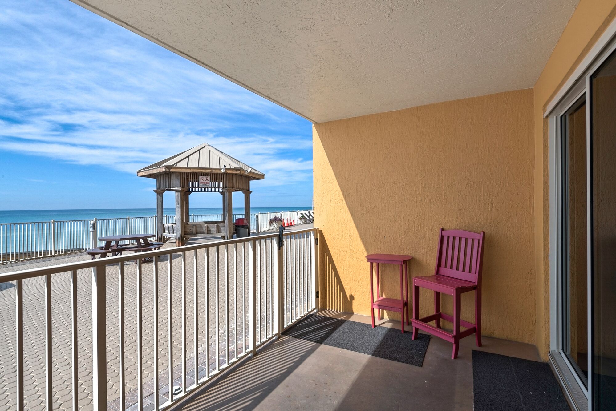 Un balcon avec vue sur la plage, un kiosque en bois et deux chaises roses.