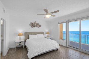 Une chambre avec un lit, deux tables de chevet, un ventilateur de plafond, un élément de décoration murale et une grande fenêtre donnant sur l’océan.