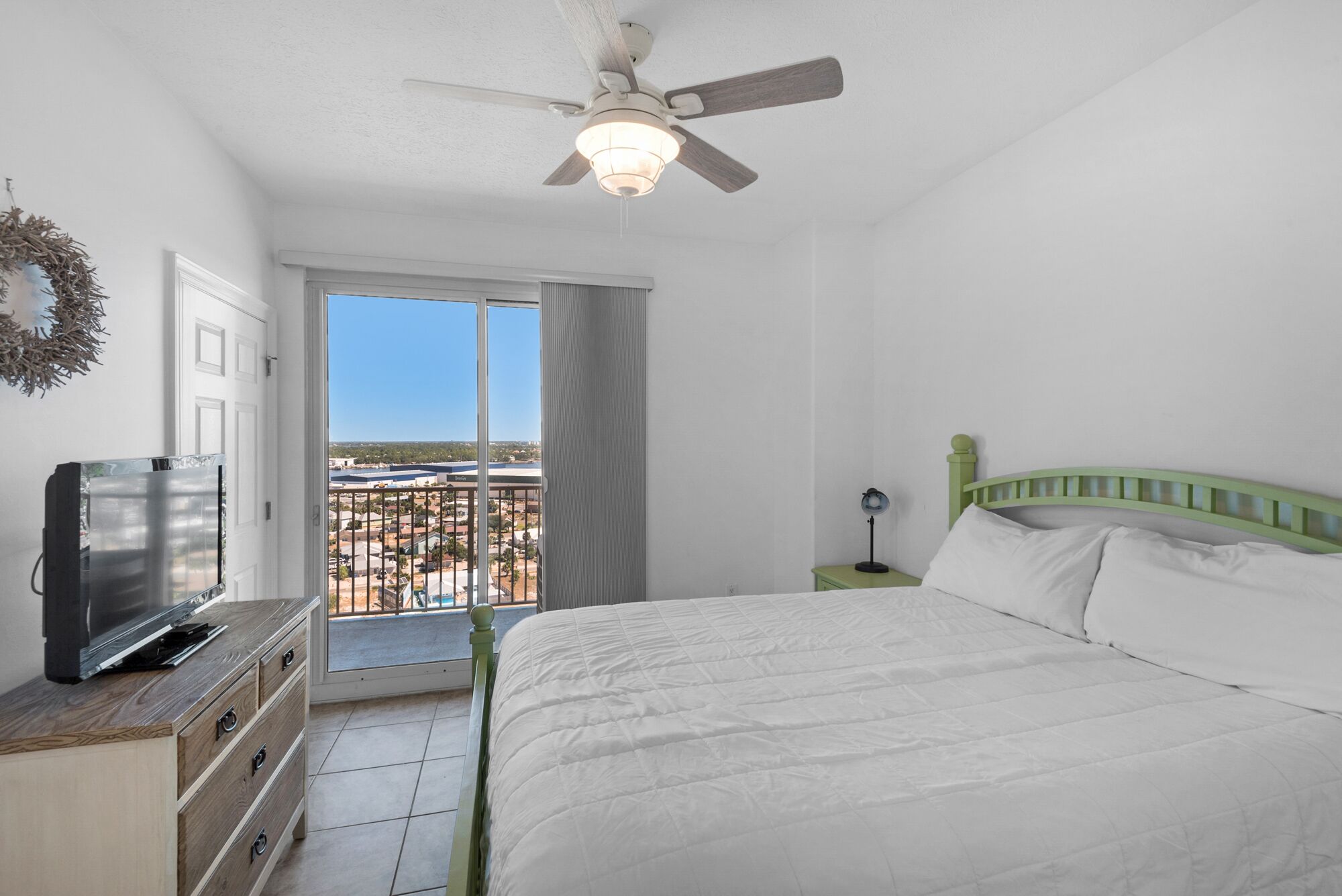 A bedroom with a bed, a ceiling fan, a TV, and a view of a residential area.