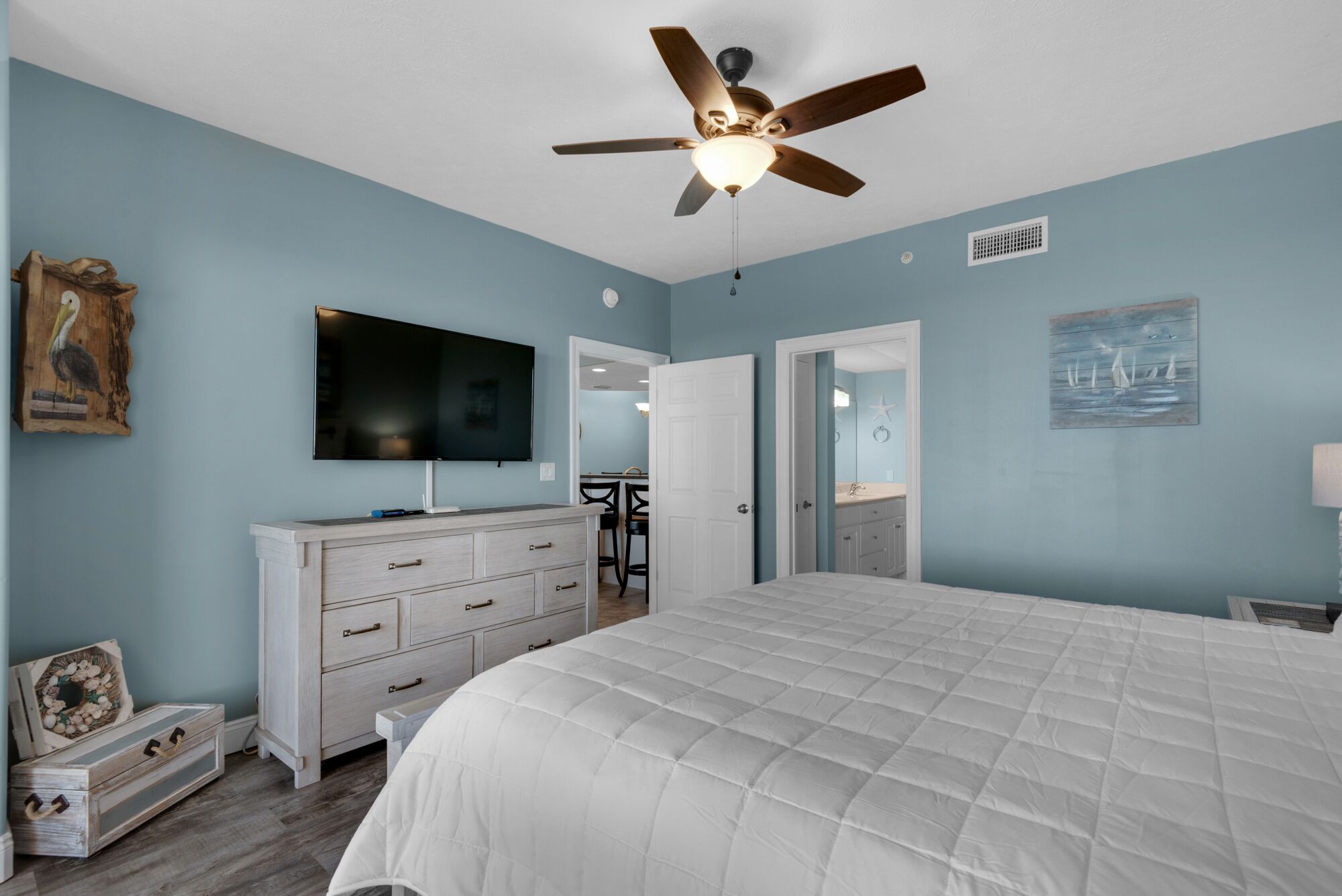 A bedroom with a bed, a dresser, a television, a ceiling fan, and artwork on the walls.