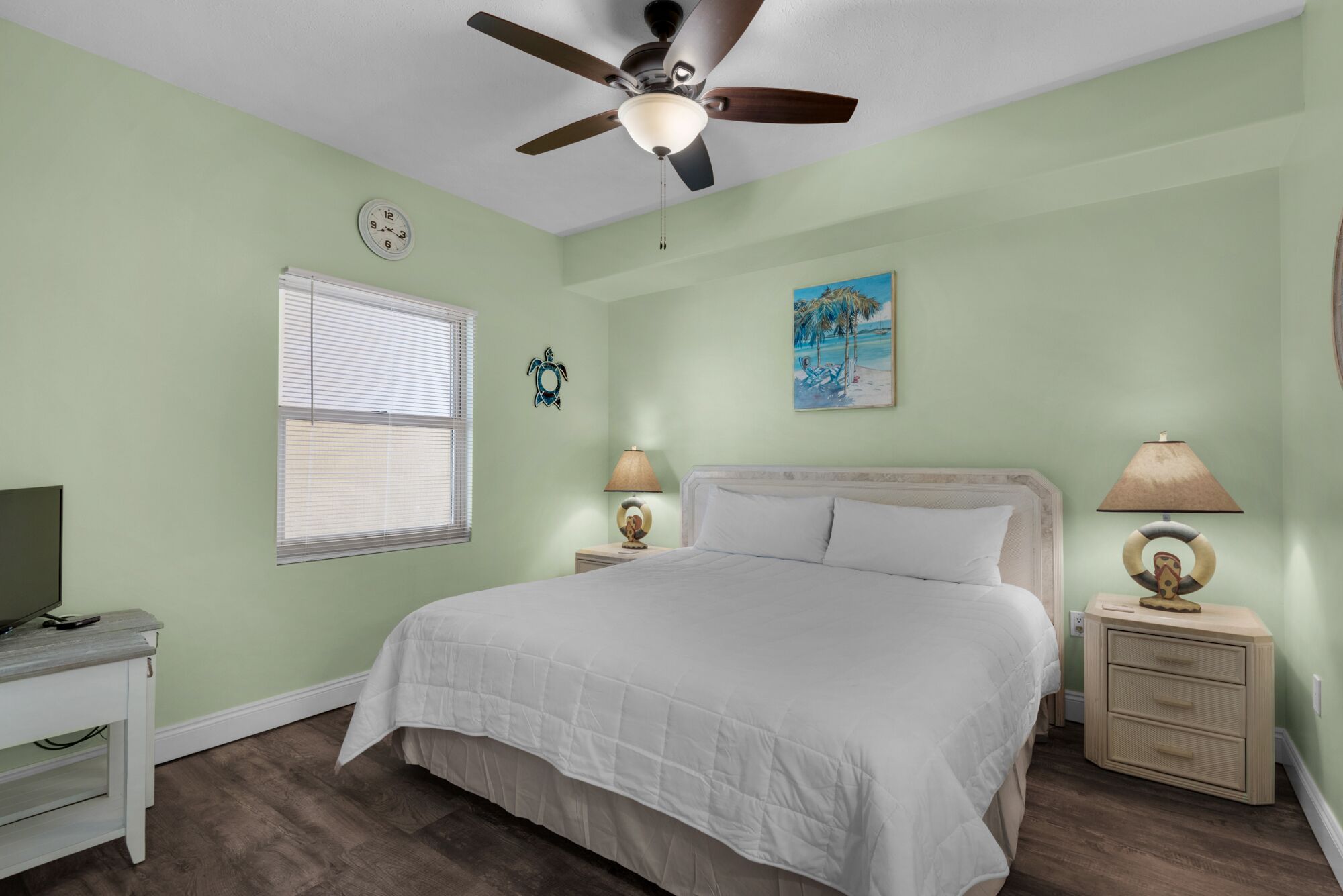A bedroom with a bed, a ceiling fan, a clock, a window with blinds, a painting, two bedside lamps, and a nightstand.