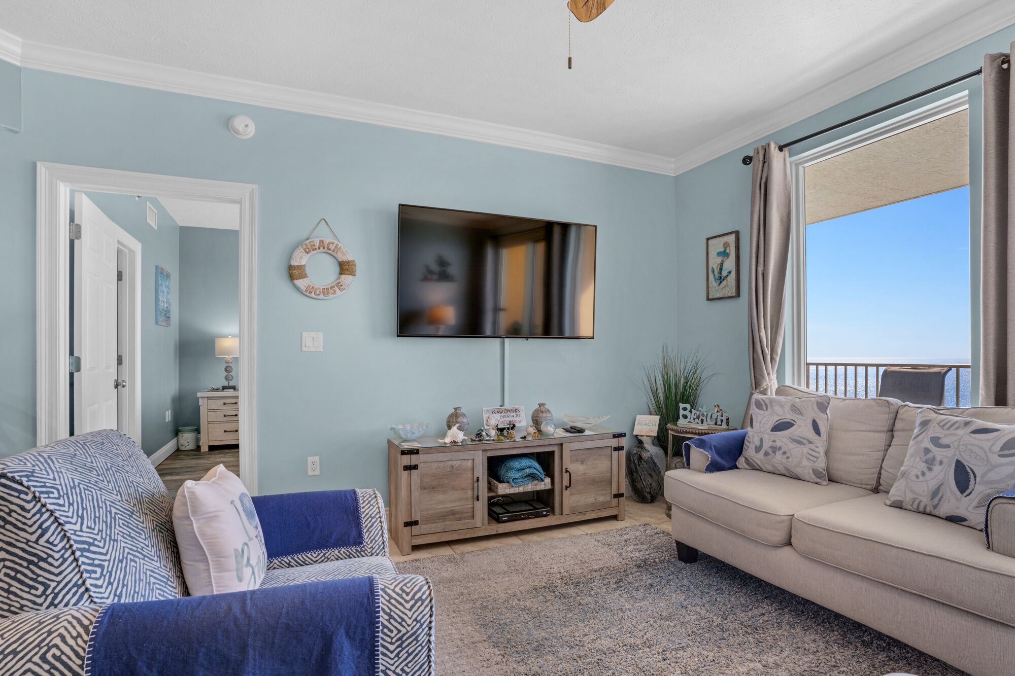 A living room with a beige sofa, a blue patterned armchair, a wooden entertainment center, and a large TV on the wall.