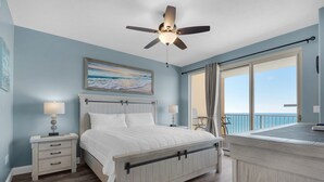 A bedroom with a bed, nightstands, a ceiling fan, a painting, and a view of the ocean. - Treasure Island 1904 (Panama City Beach)