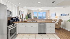 Une cuisine moderne avec des meubles blancs, un lave-vaisselle, une cuisinière et une grande fenêtre donnant sur l’océan.