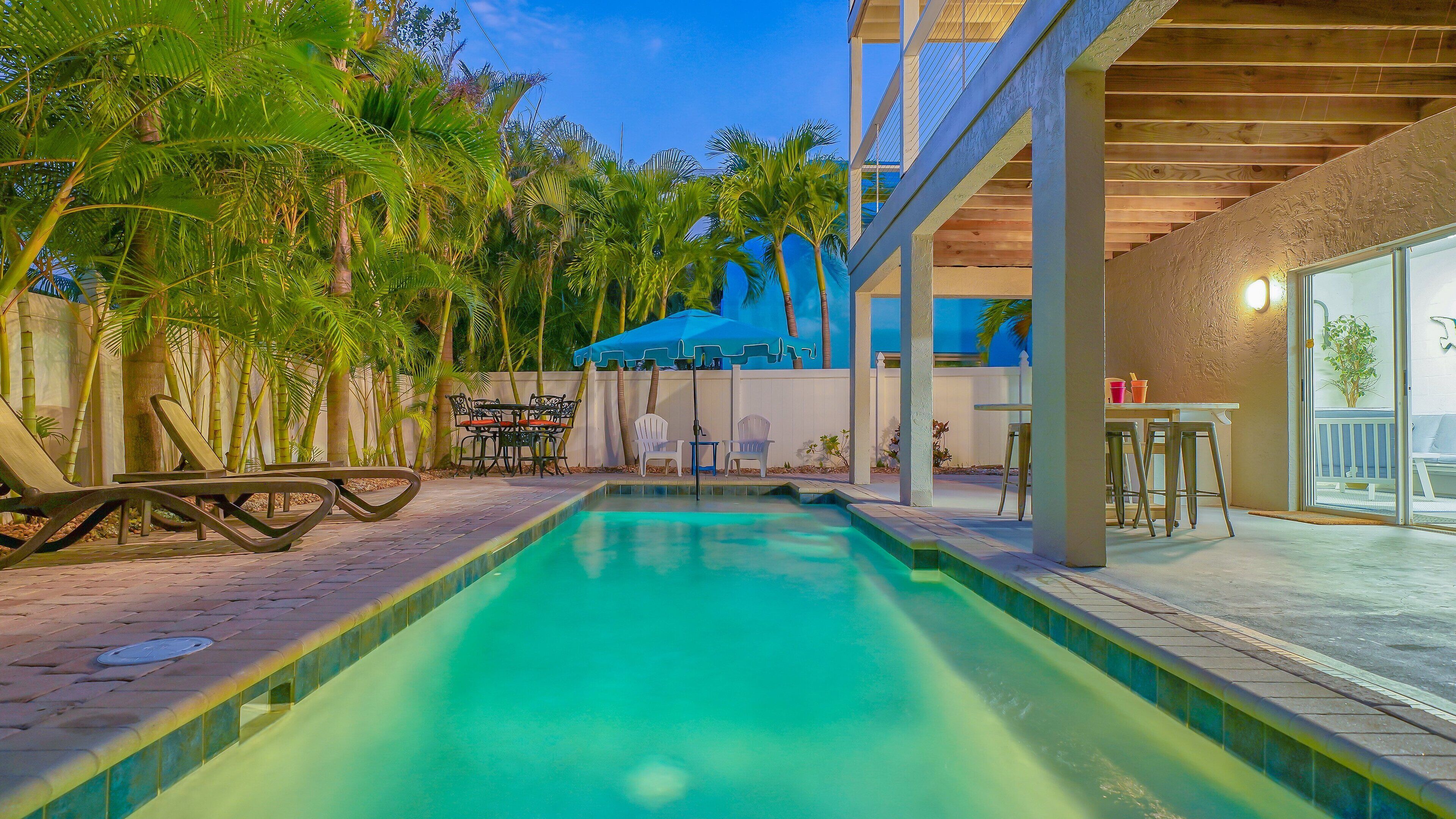 A swimming pool with lounge chairs and a table, surrounded by palm trees and a covered patio area.