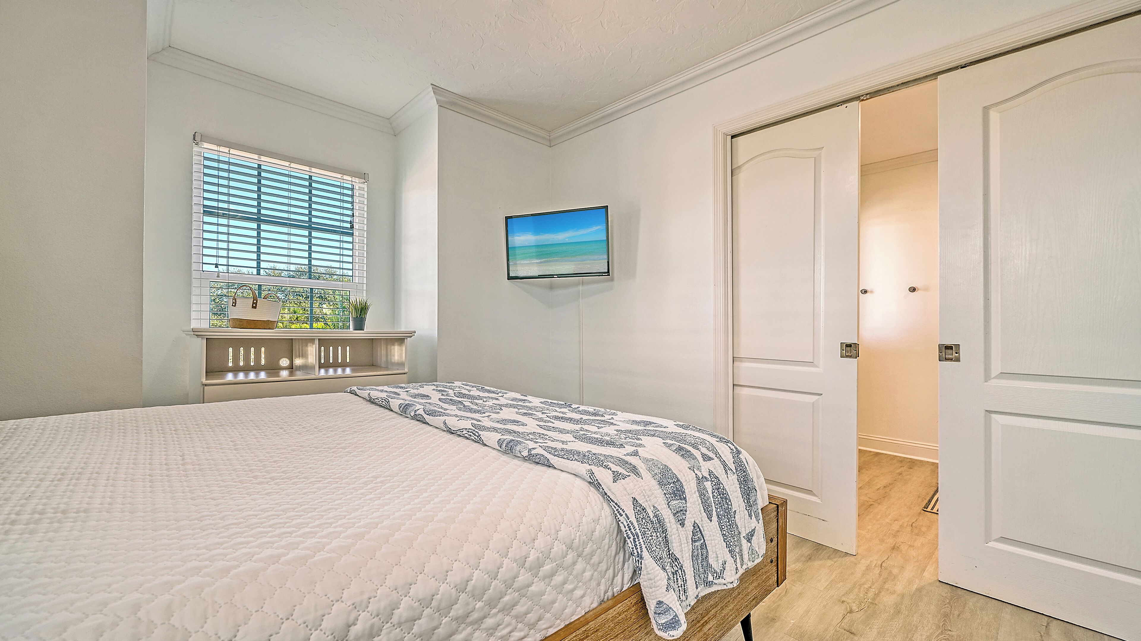 A bedroom with a bed, a window with blinds, a mounted TV, and a door.