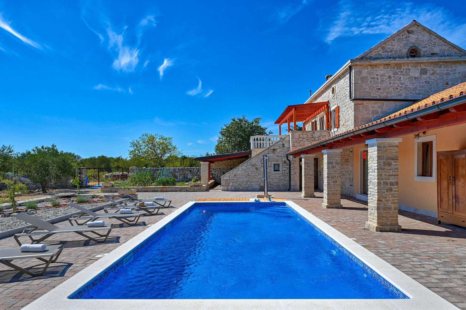 A swimming pool with lounge chairs, a stone building with a red roof, and a clear blue sky.