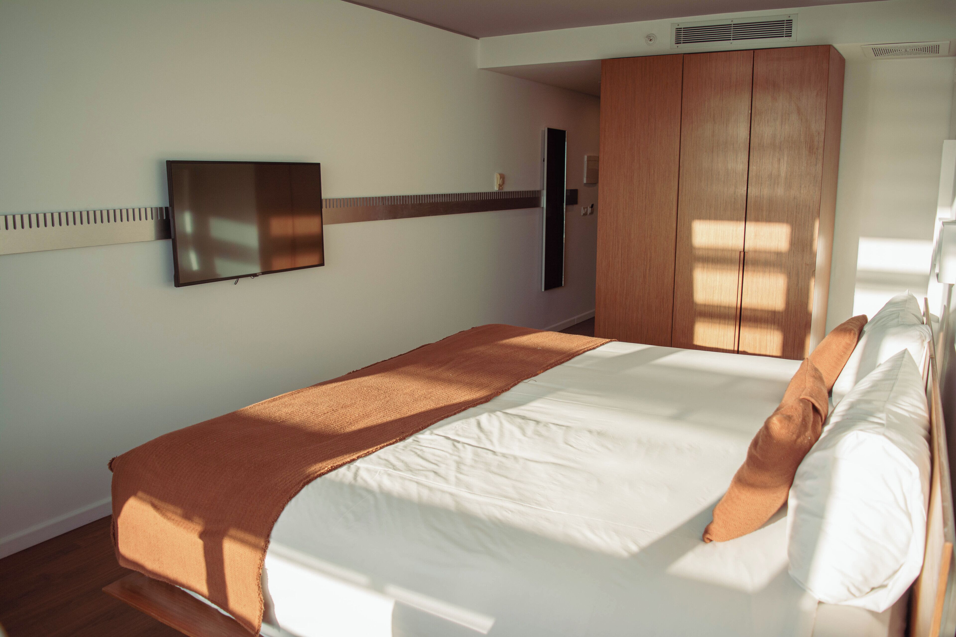 A neatly made bed with a brown blanket, a flat-screen TV mounted on the wall, and a wooden wardrobe.