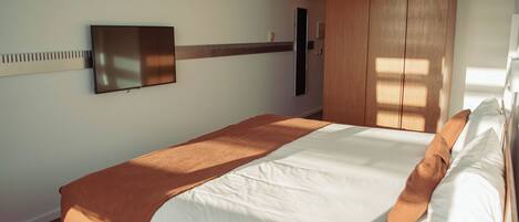 A neatly made bed with a brown blanket, a flat-screen TV mounted on the wall, and a wooden wardrobe.