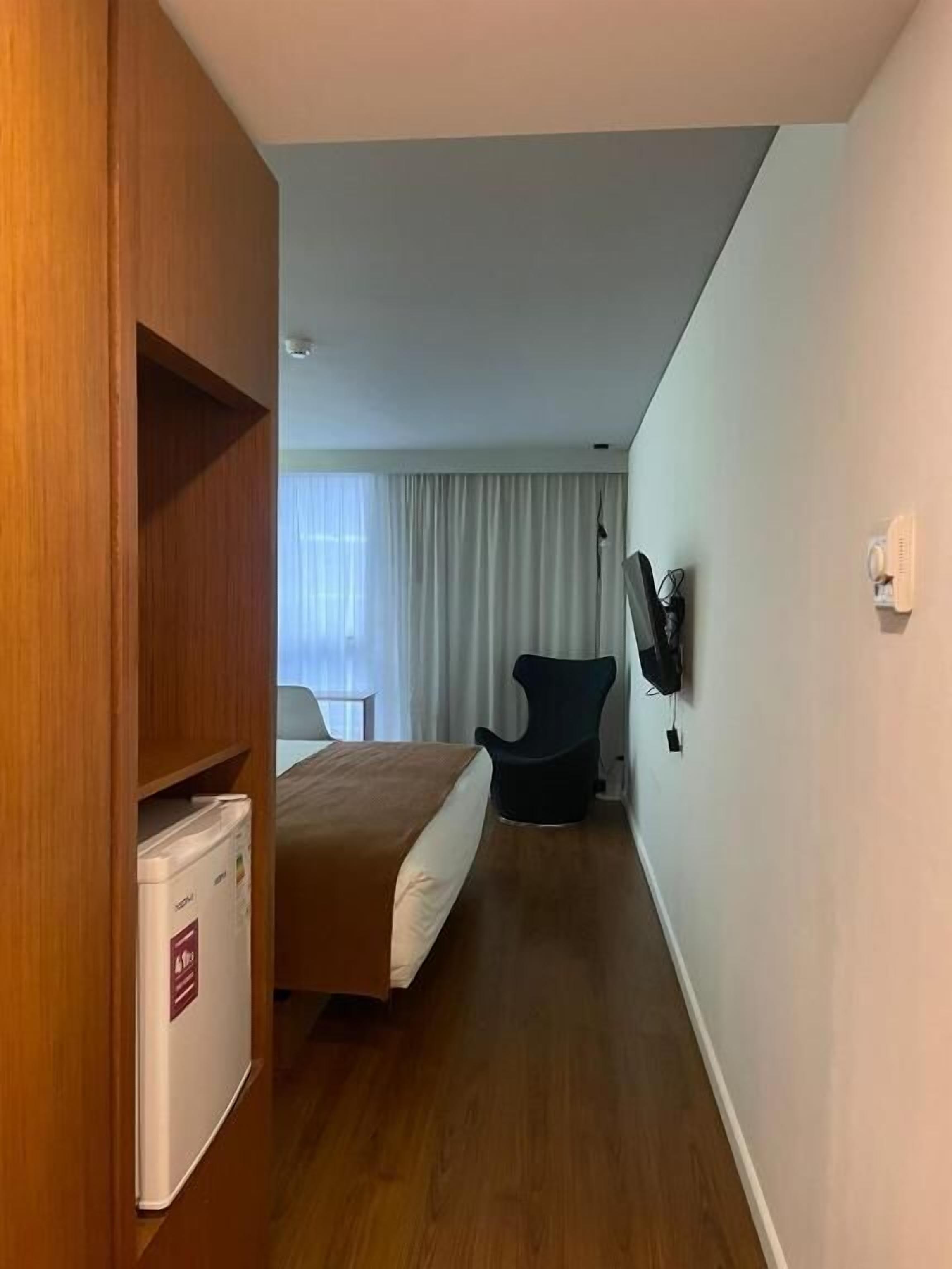 A hotel room with a bed, a chair, a TV mounted on the wall, and a minibar.