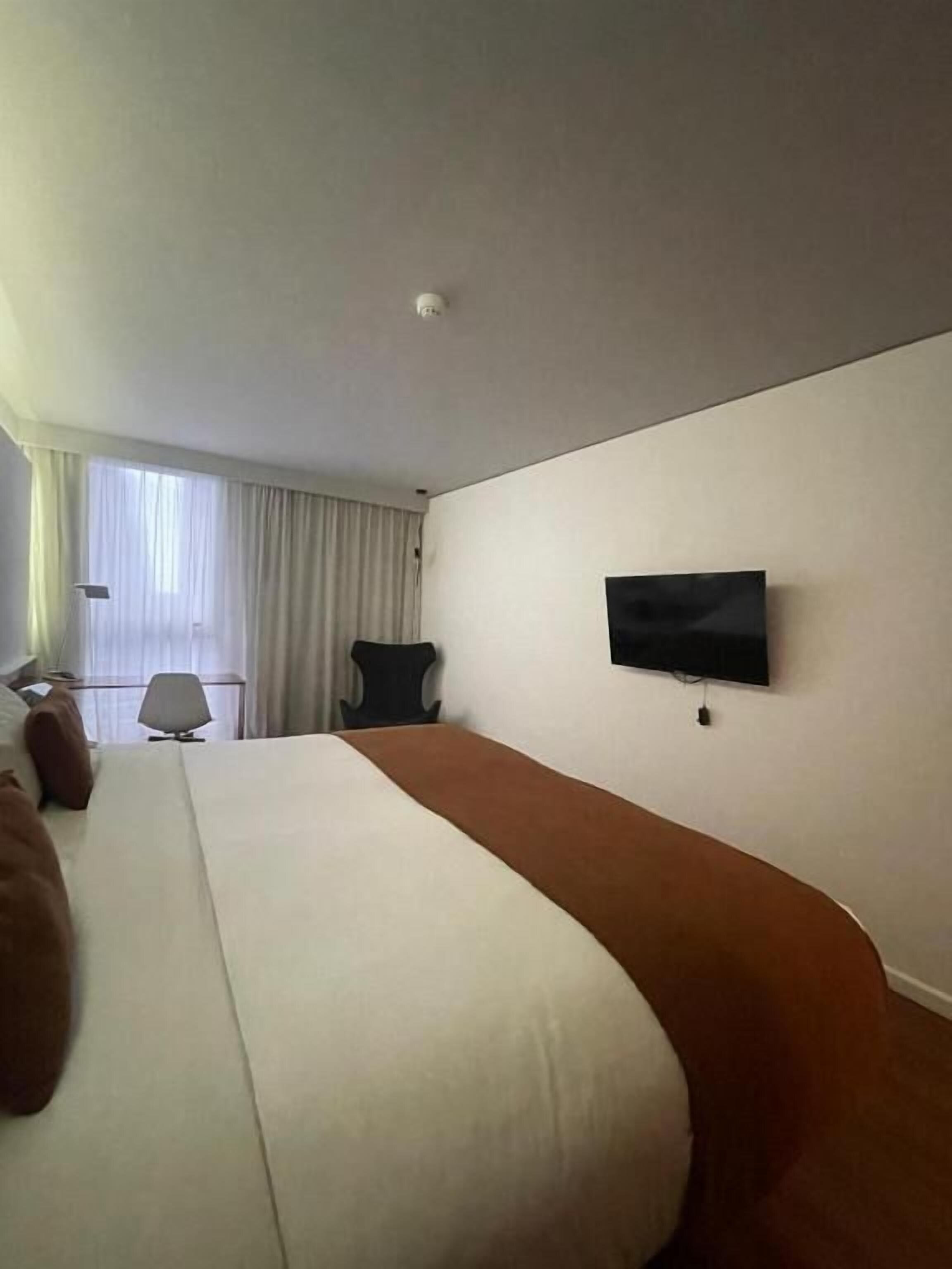 A hotel room with a large bed, a TV mounted on the wall, a chair, and a window with curtains.
