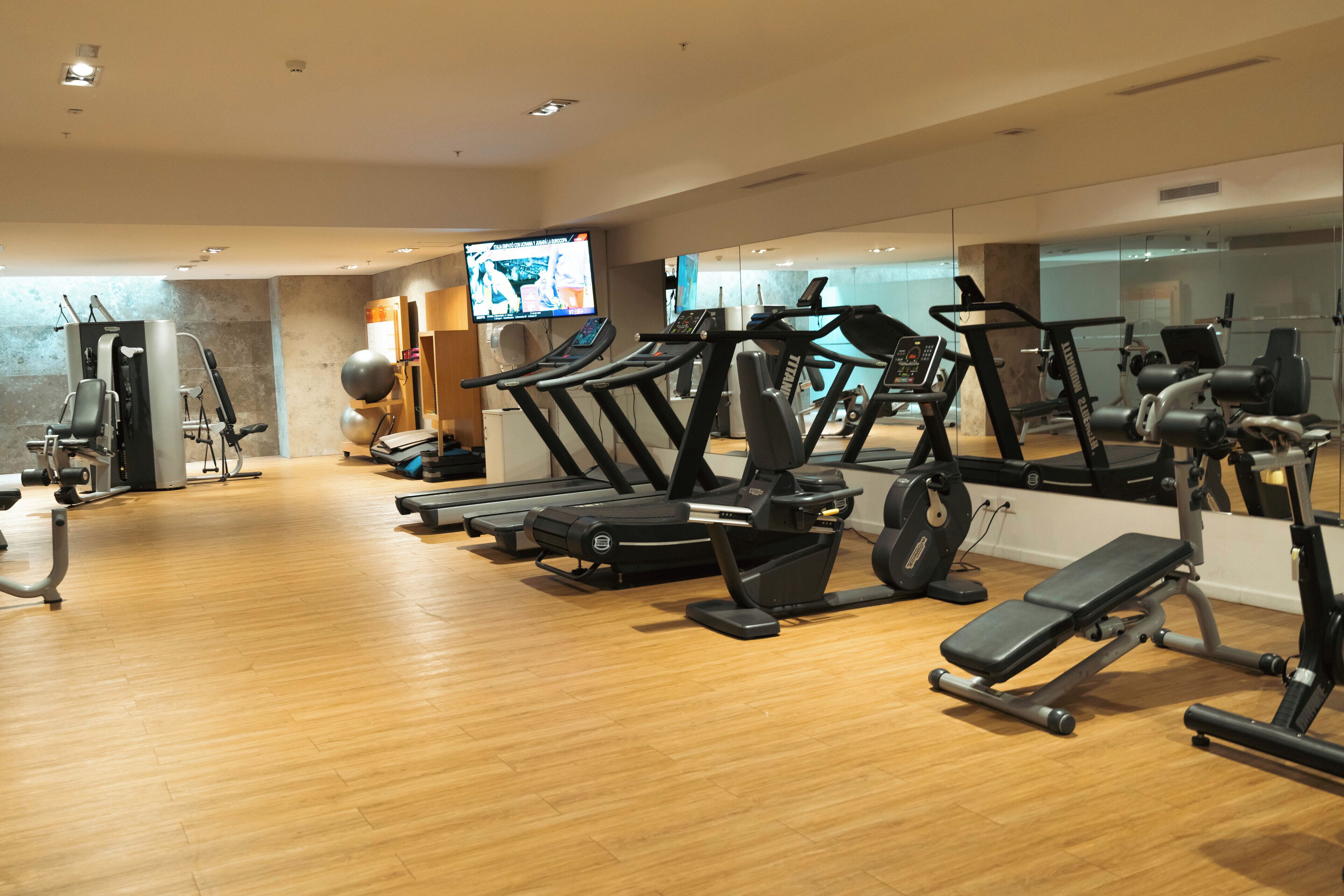 Un gimnasio moderno con varios equipos de ejercicio, un televisor mostrando videos de entrenamiento y piso de madera.