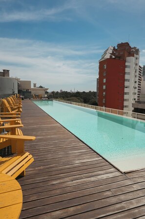 A rooftop pool with wooden deck and yellow chairs, surrounded by buildings.