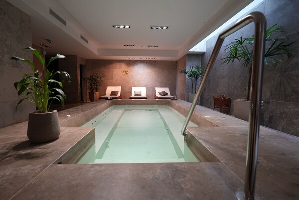 A modern indoor pool area with three lounge chairs, a potted plant, and a metal handrail.