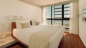 A modern hotel room with a large bed, a bedside table, a lamp, and a view of the sea through a large window. - Opta Coliving (Montevideo)