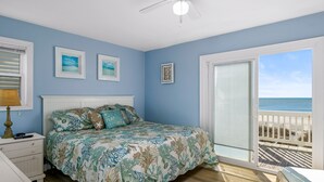 A bedroom with a bed, a nightstand, a lamp, and a view of the ocean through a sliding glass door.