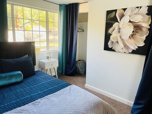 Un dormitorio con cama, mesita de noche, ventana con cortinas, un cuadro y un bolso en el suelo.