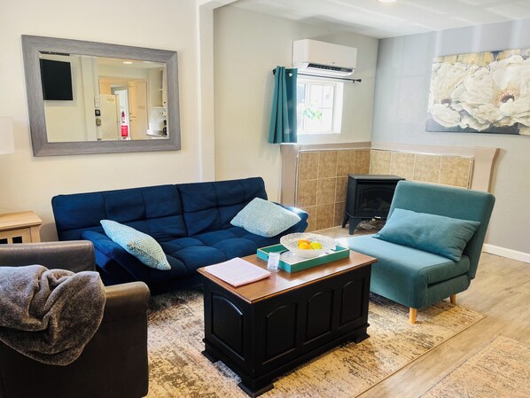 A living room with a blue sofa, a green armchair, a wooden coffee table, a fireplace, and a large mirror on the wall. - Dafna, Unit 4 - One Bedroom, Cozy Little Home (Cromberg)