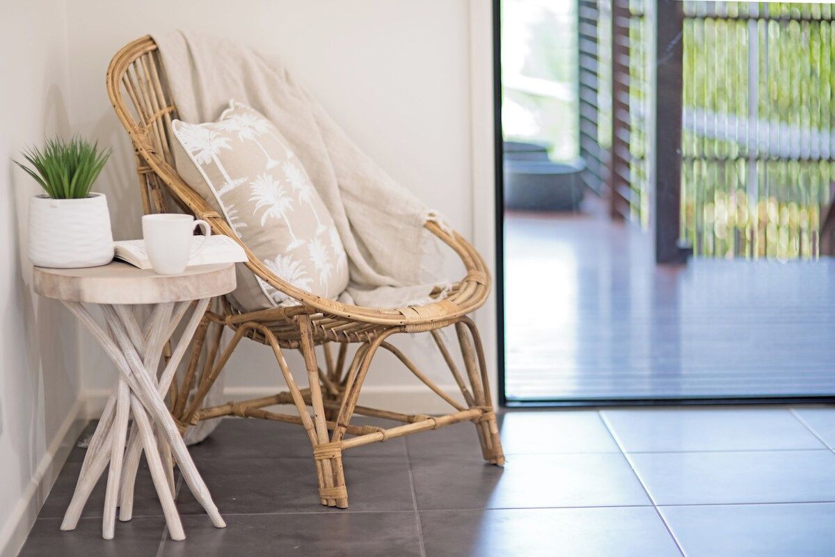 A wicker chair with a cushion, a small table with a potted plant and a cup, and a tiled floor.