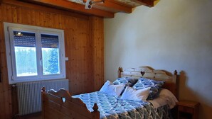 Uma cama com estrutura de madeira, colcha e almofadas estampadas, uma pequena mesa de cabeceira e uma janela com persianas.