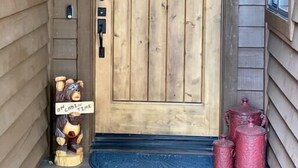Une porte en bois ornée d’un motif quadrillé, une petite figurine d’ours avec une pancarte, et deux récipients rouges sur le porche.