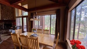Un coin repas avec une table et des chaises en bois, une cheminée en pierre et de grandes fenêtres donnant sur la forêt.