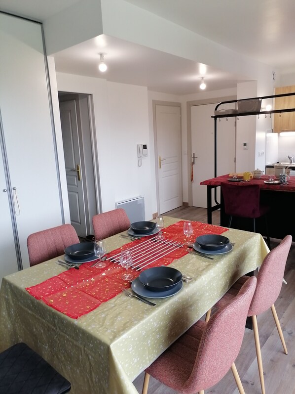 Un comedor con una mesa dispuesta para cuatro personas, incluyendo platos, cubiertos y vasos. Hay sillas alrededor de la mesa, y se distingue una zona de cocina al fondo.