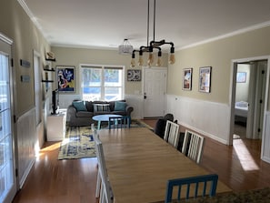 A spacious living room with a wooden dining table, a sofa, and a TV. There are large windows and a door leading to another room.