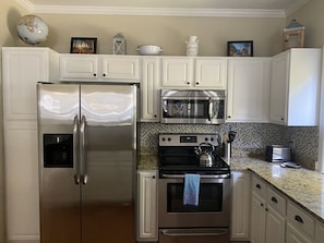 A kitchen with white cabinets, a stainless steel refrigerator, a microwave, an oven, and a kettle.