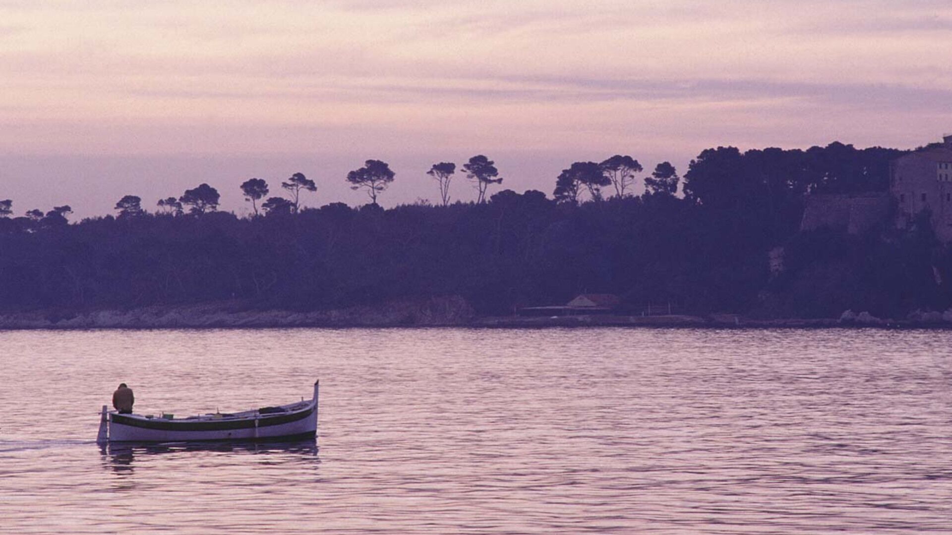 A calm river with a single boat carrying a person. The riverbank is lined with trees and a building.