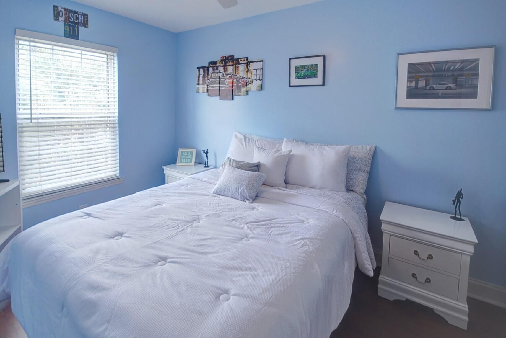 A bedroom with a large bed, a nightstand with drawers, a window with blinds, and wall art including a framed picture and a car-themed collage.