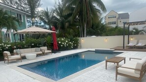 Una piscina con sillones de descanso y una zona de estar cubierta, rodeada de plantas tropicales y palmeras.