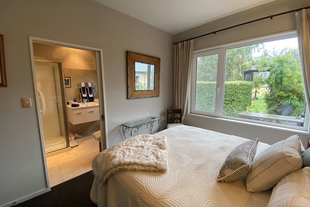 A bedroom with a bed, a bedside table, a window with a view of greenery, and a bathroom visible through an open door.