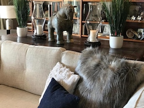 A living room with a beige sofa, a dark blue pillow, and a shaggy throw. There are two potted plants, a glass vase, and a metal sculpture on the coffee table. - Chateau Pritchard (Springston)
