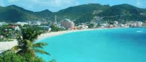 A coastal town with a beach, clear blue waters, and green hills in the background.