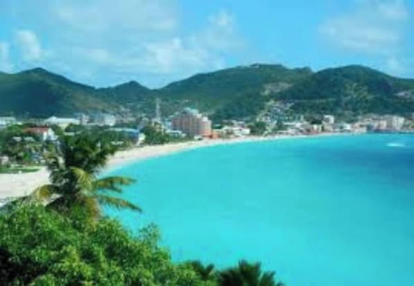 A coastal town with a beach, clear blue waters, and green hills in the background.