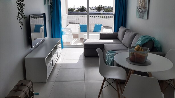 A modern living room with a sofa, a dining table, and a TV. There is a balcony with blue curtains and a view of the outside. - MARINE SENATOR (Costa Teguise)
