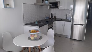 A modern kitchen with white cabinets, a stainless steel refrigerator, a microwave, and a sink. There is a round table with chairs and a basket of fruit. - MARINE SENATOR (Costa Teguise)