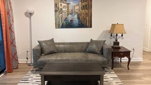 A living room with a sofa, coffee table, floor lamp, and a painting of a canal scene. - 2144 New Bayfront Condo overlooking Mobile Bay (Daphne)