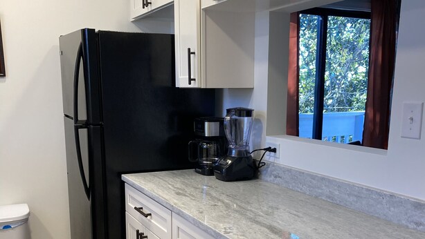 A modern kitchen with white cabinets, a black refrigerator, a coffee maker, and a window with curtains. - 2144 New Bayfront Condo overlooking Mobile Bay (Daphne)