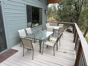 A covered outdoor patio with a glass table and chairs, a wooden deck, and a view of trees.