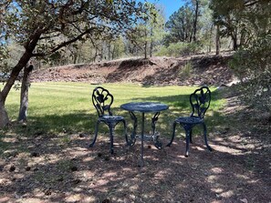 An outdoor seating area with two metal chairs and a round table, surrounded by trees and a grassy field.