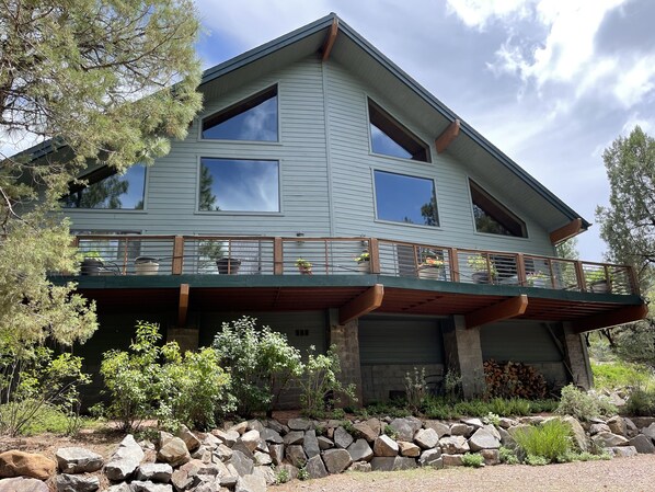 A two-story house with a balcony, stone foundation, and surrounding greenery.