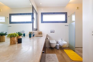 A modern bathroom with a large sink, toilet, bidet, and a mirror. There are potted plants on the counter and a yellow rug on the floor. - Villa-4bedroom-3bathroom-pool-petfriendly (Nazaré)