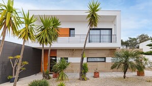 A modern two-story house with a balcony, surrounded by various potted plants and palm trees. - Villa-4bedroom-3bathroom-pool-petfriendly (Nazaré)