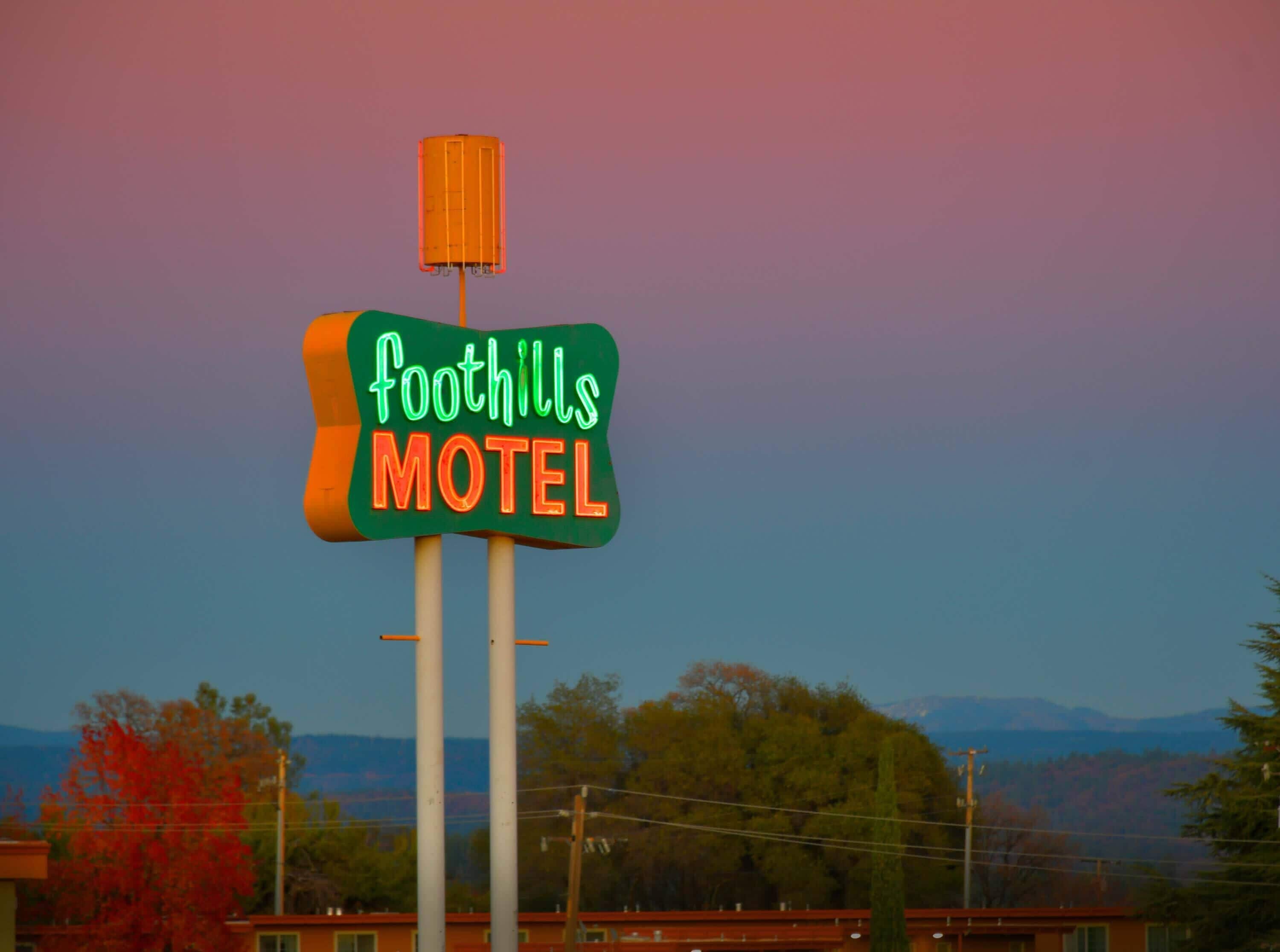 Une enseigne de motel portant le nom « Foothills Motel », avec en arrière-plan des arbres aux couleurs automnales et des montagnes lointaines.