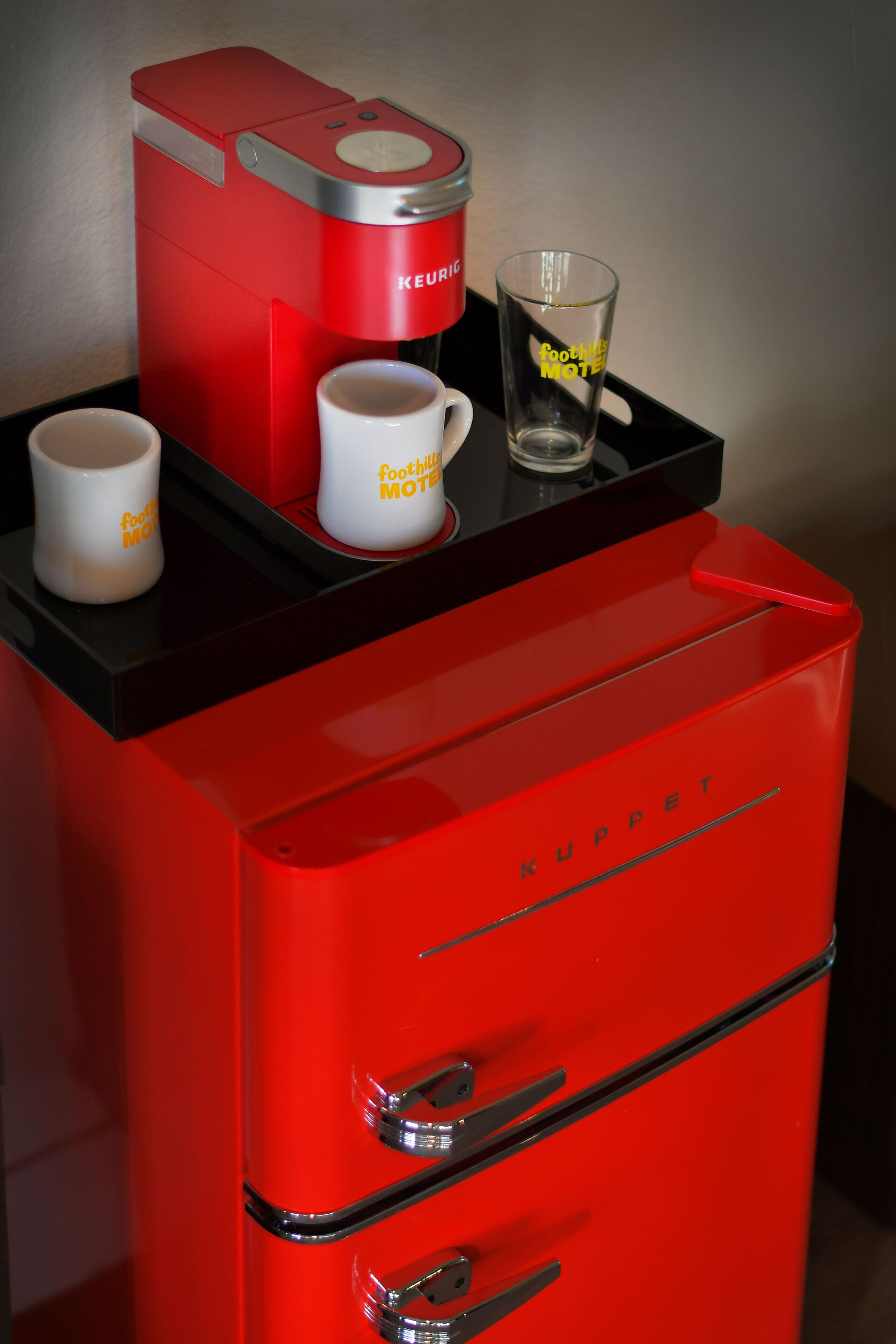 A red coffee machine with two white mugs and a glass on a black tray.