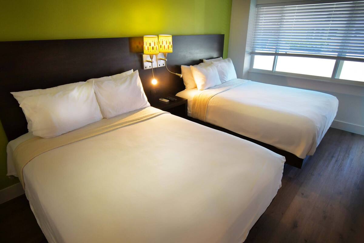 A hotel room with two beds, a nightstand with a lamp, and a window with blinds.